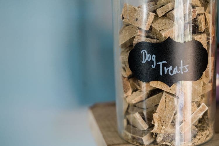 A close-up image of dry dog treats stored in a labeled glass jar on a wooden surface.